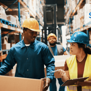 Group of warehouse workers in hard hats and safety gear, carrying boxes and clipboards